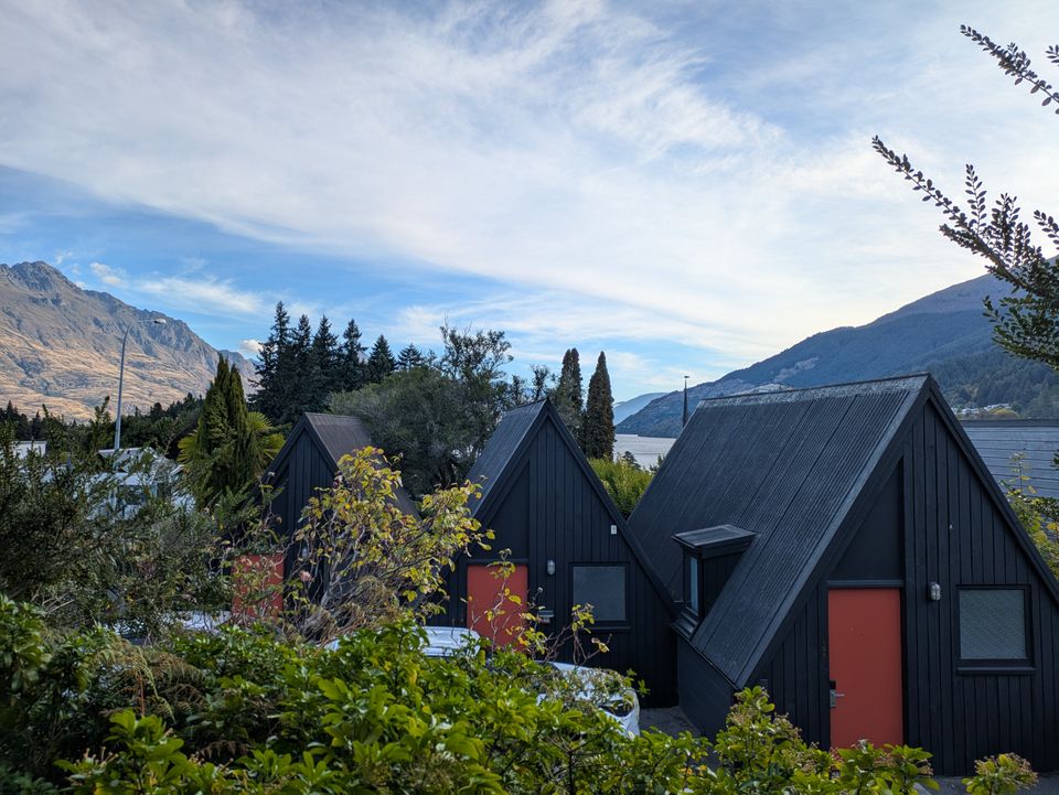 A row of black A-frame cabins with red doors against snow-streaked mountains and a lake at golden hour