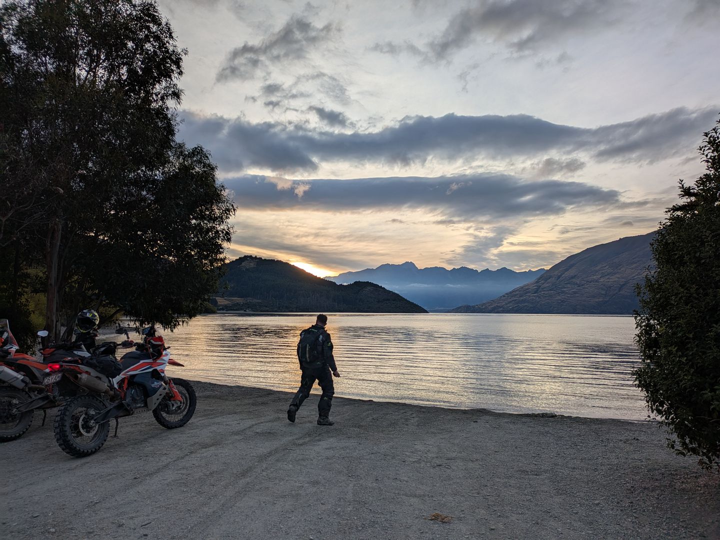 Two KTMs parked at the edge of a lake at dusk, a rider walking toward the water under mountain silhouettes and a long pastel sky