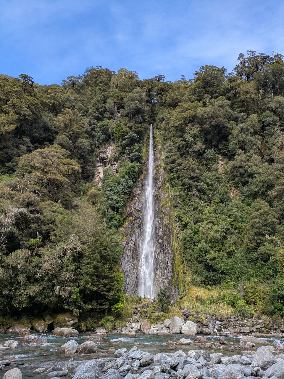 A tall narrow white waterfall cutting through a forested cliff into a rocky river