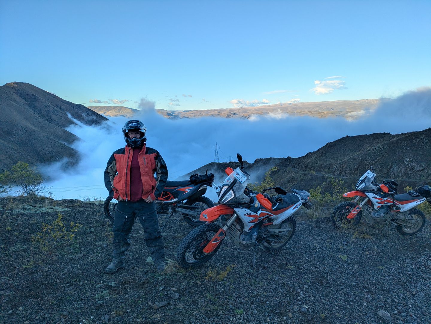 A rider in riding gear standing on a ridge next to two KTM motorcycles, looking out over a valley filled with low cloud at sunrise