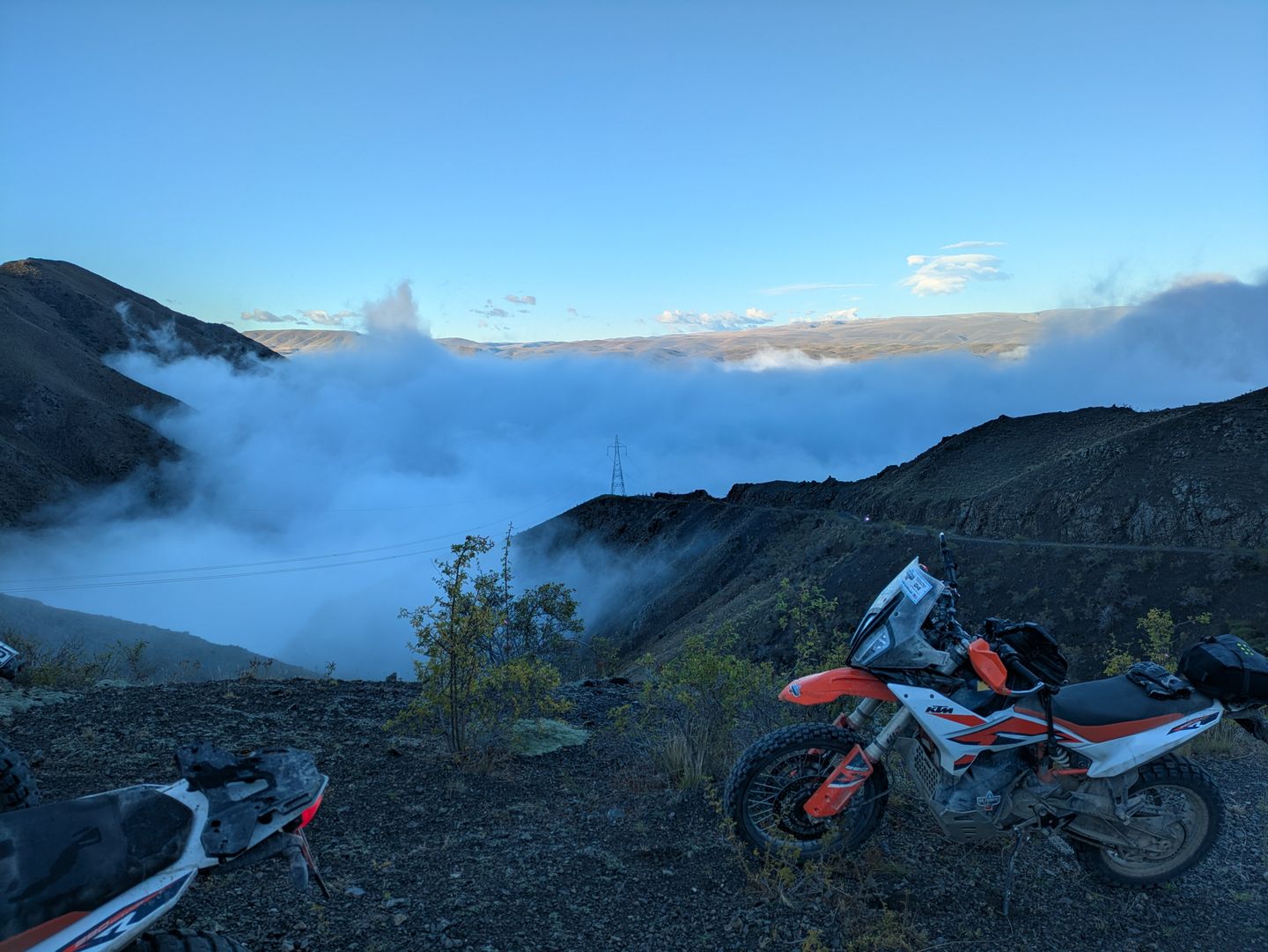 A KTM adventure motorcycle on a high ridgeline above a cloud-filled valley at sunrise, New Zealand South Island